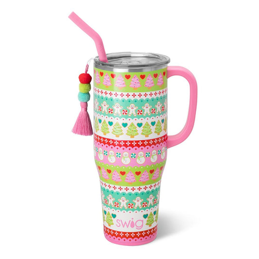 A pink handled travel tumbler with a stainless steel lid, pink straw, and a decorative tassel charm. The tumbler features a colorful Christmas-themed pattern with gingerbread men, snowmen, candy canes, and Christmas trees in stripes of green, teal, red, and white.